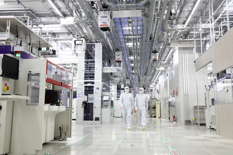 Interior of a Samsung semiconductor fabrication plant cleanroom, with engineers in full protective suits walking between manufacturing equipment